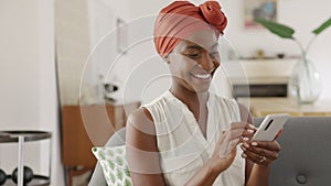 Smiling black woman with african turban using smartphone at home