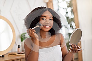 Smiling black woman applying cosmetic powder or blusher on her face, using decorative makeup, looking in mirror at home