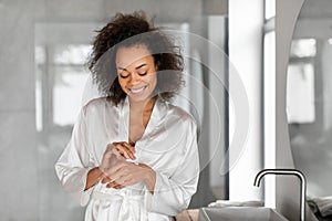 Smiling Black Woman Applying Moisturizer Cream On Hands In Bathroom