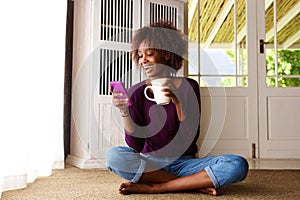 Smiling black woman sitting on floor at home with cell phone