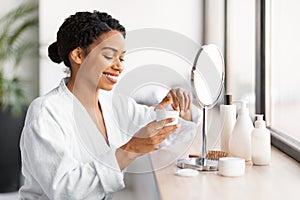 Smiling black woman in white bathrobe opening jar of facial cream