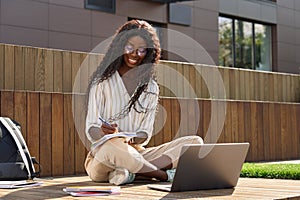Happy pretty African girl student studying online using laptop sitting outside.