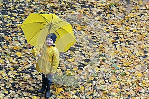Smiling blod boy with large yellow umbrella and raincoat on a background of yellow autumn foliage. Top view