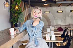 Smiling blond girl sitting in a coffee shop drinking coffee and touching her hair. She wears blue denim shirt and grey