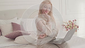 Smiling blond grey-eyed Caucasian businesswoman sitting with laptop on the bed and thinking. Young beautiful lady in