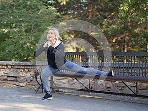 Smiling blond grownup woman in hoodie doing one leg squat using a bench in urban park in summer, outdoor, selective