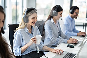 Smiling blond haired woman operator with headset drinking morning coffee and starting doing her online job at customer support