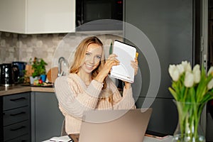 Smiling blond young woman with worksheets studying online, e-learning using laptop at home. Remote distance study