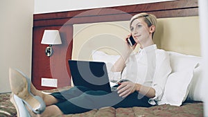 Smiling businesswoman in white shirt using on laptop and talking at mobile phone while lying on bed in hotel room