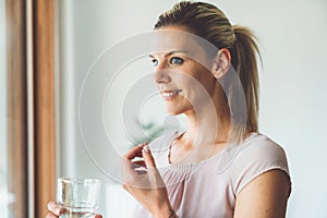 Smiling blonde caucasian woman holding a vitamin pill in one hand and a glass of water in other - Taking supplements