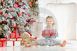 Smiling blonde girl holding christmas gift box. Child 5-6 years old playing by fireplace in festive decorated room. Celebrating