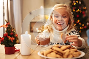 Smiling blonde girl holding mug with cacao and cookie, looking a