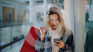 Smiling blonde girl reads something in her phone walking with shopping bags around a shopping mall