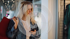 Smiling blonde girl reads something in her phone walking with shopping bags around a shopping mall