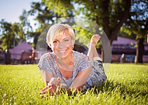 A smiling blonde girl with a short haircut is lying in a city park on green grass. A woman is resting in a park and looking into