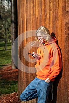 Smiling blonde hipster guy reads funny message in phone chats with friends stands near wooden fence.