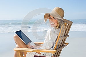 Smiling blonde sitting on wooden deck chair by the sea using tablet