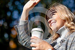 Smiling blonde in tweed jacket with cup of hot drink in his hands, he squints, closing his hand from direct sunlight