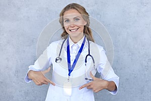 Smiling blonde woman doctor wearing uniform standing isolated over grey background, showing her name on badge