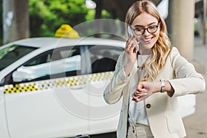 smiling blonde woman in eyeglasses talking by smartphone and checking wristwatch while standing near taxi cab
