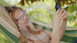 Smiling blonde woman with eyeglasses using smartphone, lying relaxing on the hammock in the garden, free time concept to surf the