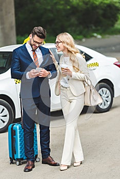 smiling blonde woman holding coffee to go and looking at handsome man using smartphone while standing with suitcase