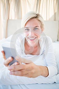 Smiling blonde woman lying on the bed and texting with her mobile phone