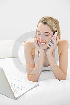 Smiling blonde woman lying on her bed phoning using her notebook