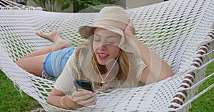 Smiling blonde woman with sunglasses using smartphone, lying relaxing on the hammock in the garden, free time and summer