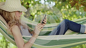 Smiling blonde woman with sunglasses using smartphone, lying relaxing on the hammock in the garden, free time and summer holiday