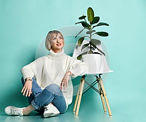 Smiling blonde woman in sweater, jeans and sneakers sits on floor at modern chair with ficus in pot on it looking up