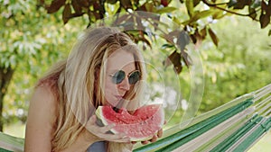 Smiling blonde woman wearing sunglasses, eating a slice of watermelon on hammock in the garden, leisure and summer holiday concept