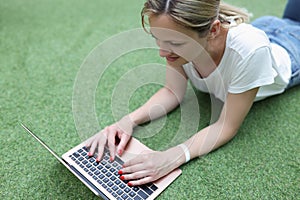 Smiling blonde woman working on laptop lay on grass, remote job, female freelancer outdoors