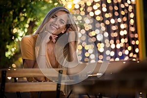 Smiling blonde young woman sitting, with evening fairy lights on the background.