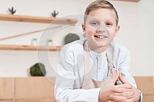 smiling boy in apron looking at camera while leaning on table