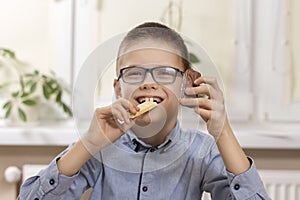 Smiling boy with glasses sits at the table and holds sweets in his hands. In one hand he holds white chocolate which he bites and