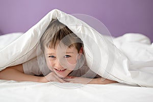 Smiling boy hiding in bed under a white blanket or coverlet
