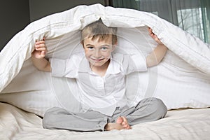 Smiling boy hiding in bed under a white blanket or coverlet.