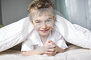 Smiling boy hiding in bed under a white blanket or coverlet.