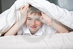 Smiling boy hiding in bed under a white blanket or coverlet.