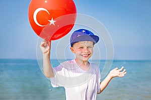 Smiling boy holds balloon with turkish flag on blue sea background on sunny summer day. Happy caucasian child on resort beach