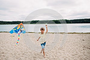 Smiling boy with a kite in his hands runs along the sandy beach on the banks of a picturesque river