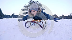 Smiling boy lying on sled rides from snow-covered hill in winter forest in slow motion