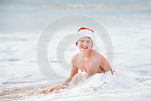 Smiling boy in Santa`s hat swims on ocean surfline. Christamas a