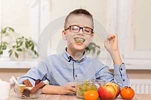 Smiling boy sits at the table and eats fruit. He has grape fruit in his teeth.