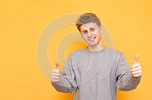 Smiling boy is standing on a yellow background, looking into the camera and showing his thumb up. Positive young man shows a