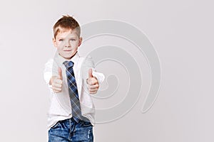 Smiling boy in white shirt and tie on grey wall background. Fashionable male child showing thumb up. Kids style fashion
