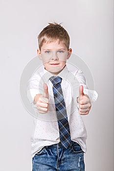 Smiling boy in white shirt and tie on grey wall background. Fashionable male child showing thumb up. Kids style fashion