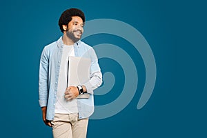 A smiling Brazilian young man, freelancer or student carrying a laptop standing isolated on blue