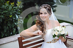 The smiling bride is dressed in a Greek-style wedding dress, her hair is covered with lalatic, holds a bouquet of white